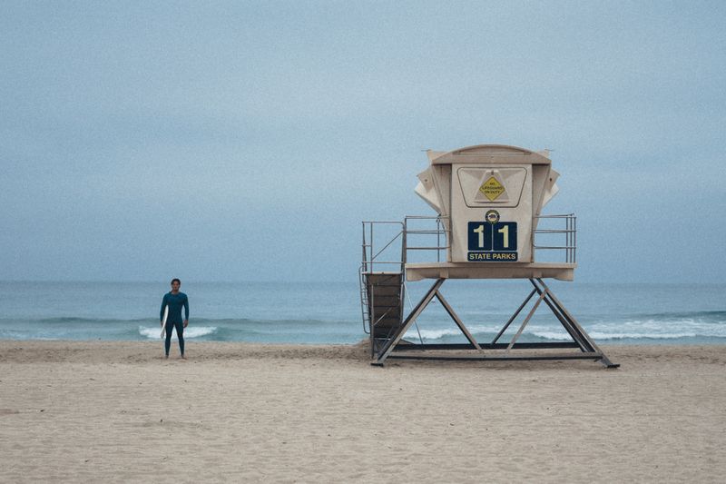 Surfer in a dark wetsuit standing on a sandy beach next to a large elevated lifeguard tower under an overcast sky.