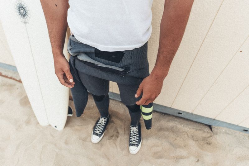 Man wearing a white t-shirt, grey wetsuit, and black sneakers standing on sand next to a surfboard.