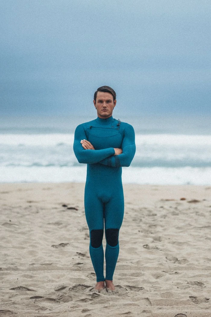 Man standing with arms crossed on a sandy beach wearing a teal wetsuit against an overcast sky and ocean horizon.