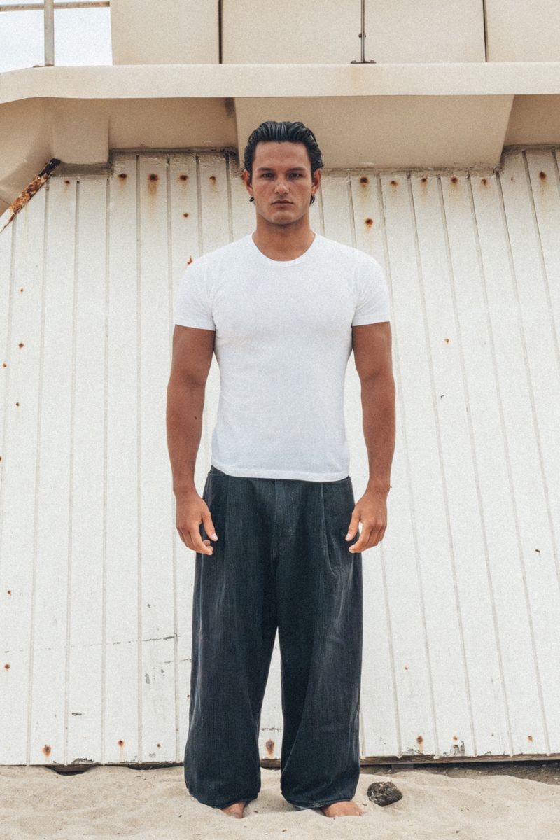 Man wearing a white t-shirt and dark trousers standing against a textured pale wall by sandy ground.