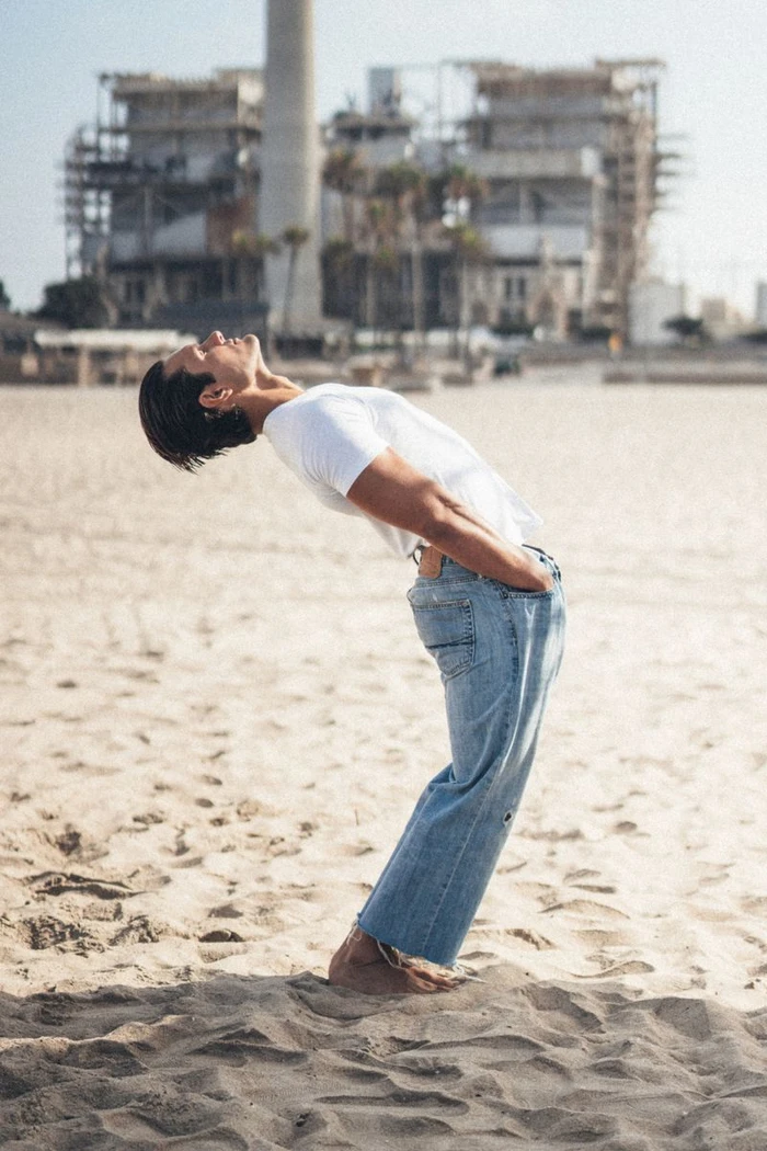 Man wearing a white t-shirt and blue jeans arching backward on a sandy beach with an industrial building in the background