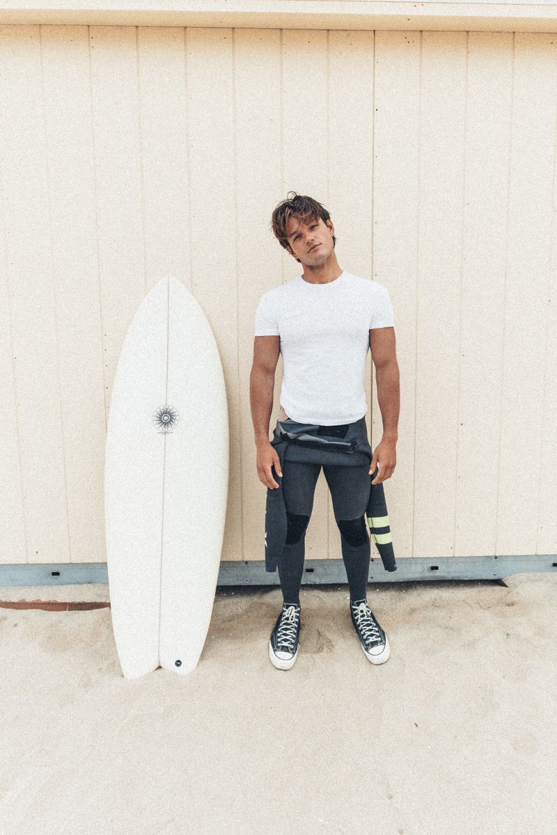 Young man standing next to a white surfboard in front of a pale wooden wall