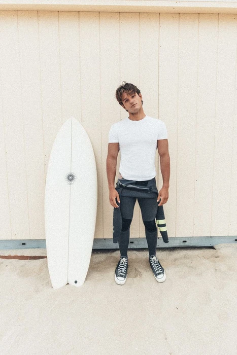 Young man standing next to a white surfboard in front of a pale wooden wall