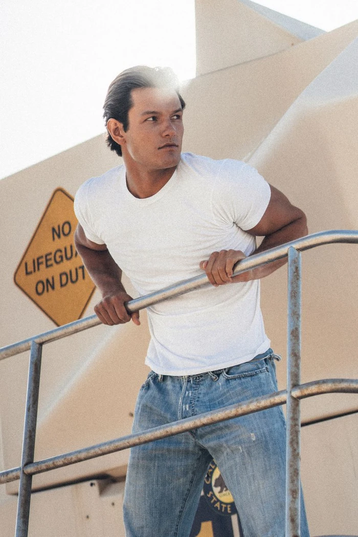 Man wearing a white t-shirt and blue jeans leaning on a metal railing with a yellow sign in the background