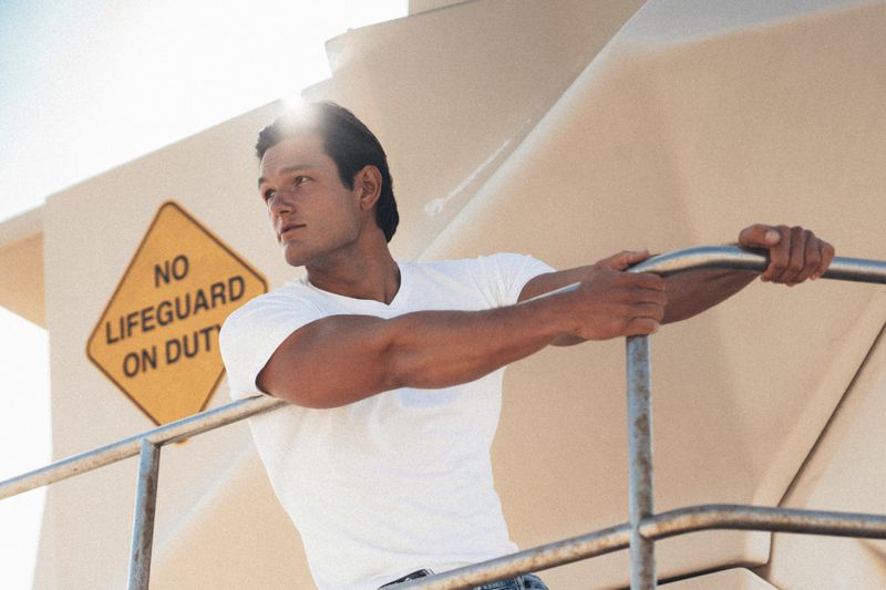 Man wearing a white t-shirt leaning against a railing with a yellow no lifeguard on duty sign in the background.
