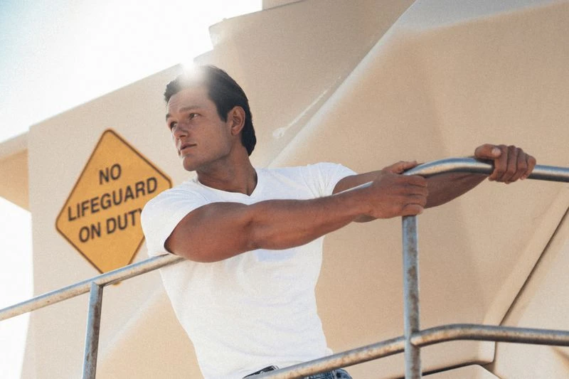 Man wearing a white t-shirt leaning against a railing with a yellow no lifeguard on duty sign in the background.