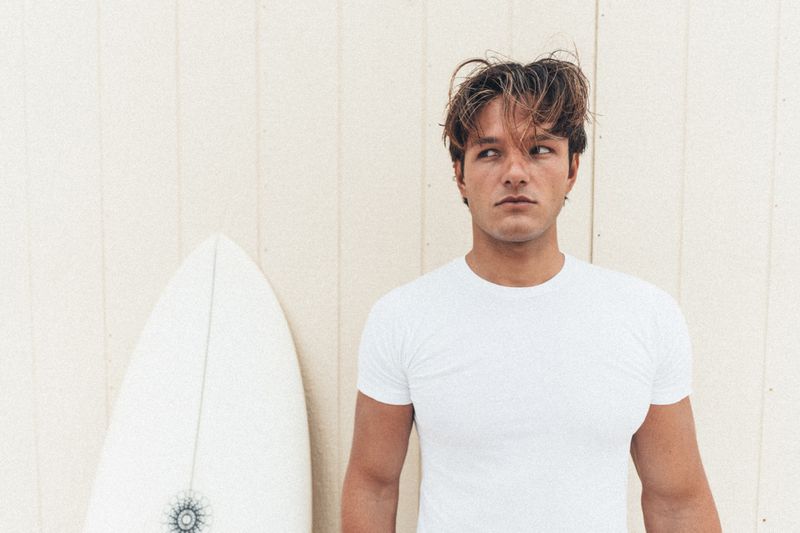 A man wearing a white t-shirt with a surfboard positioned beside him against a light wooden paneled background.