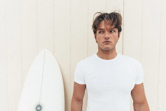 A man wearing a white t-shirt with a surfboard positioned beside him against a light wooden paneled background.