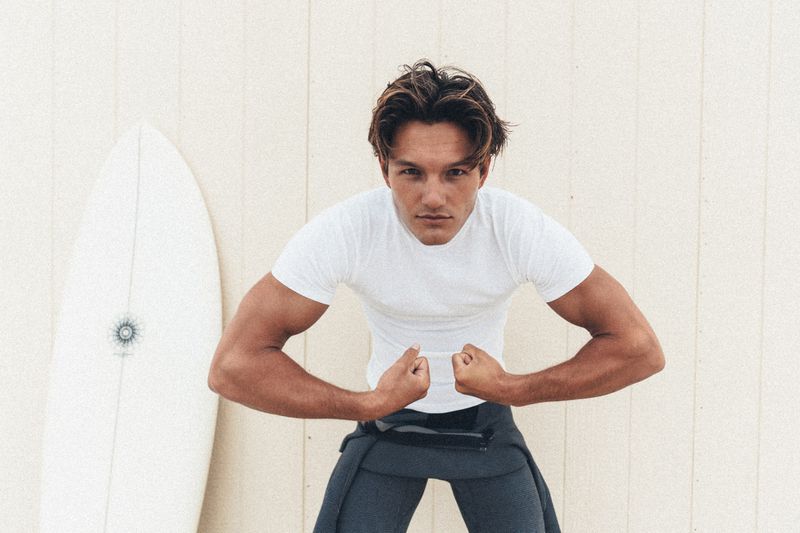 Young man in a white t-shirt and dark trousers posing with a surfboard against a light-coloured wooden wall.