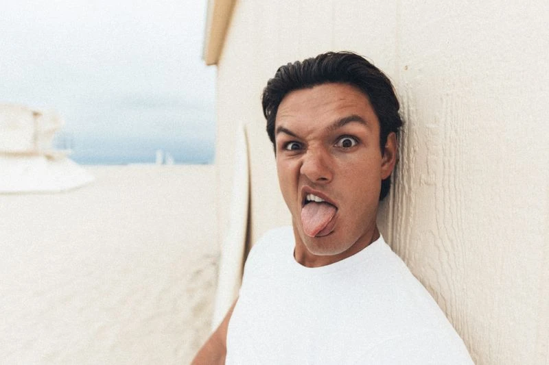 Man wearing a white t-shirt leaning against a wall outdoors, making a playful face with his tongue sticking out.