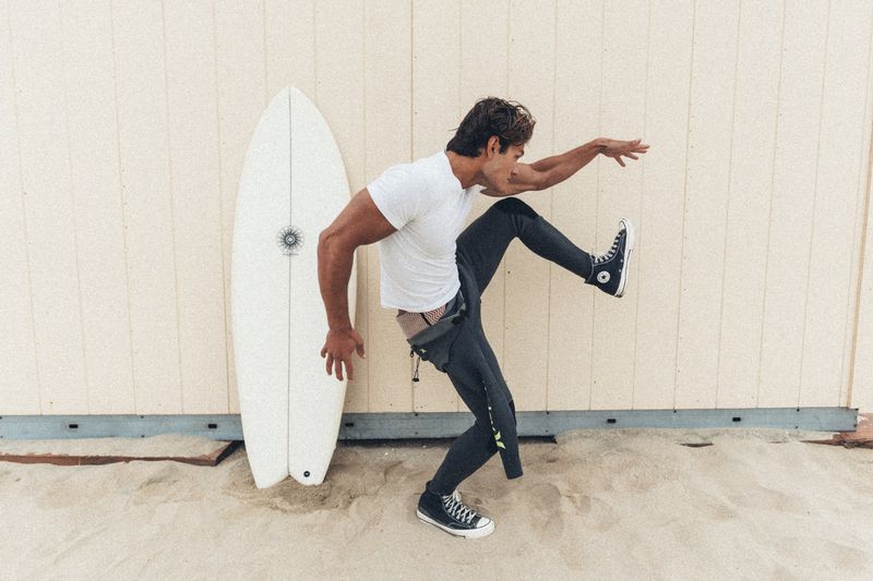 A man in a white t-shirt and wetsuit pants poses next to a white surfboard standing against a wooden wall above sand.