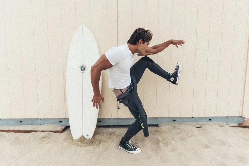 A man in a white t-shirt and wetsuit pants poses next to a white surfboard standing against a wooden wall above sand.