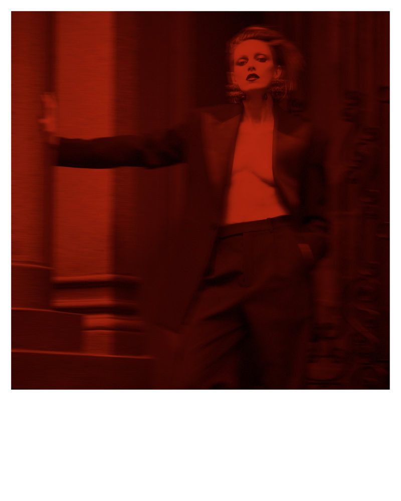 Anja Rubik in a dark blazer and trousers posed against an architectural exterior under a red monochrome tone.