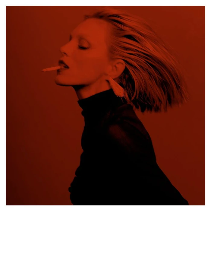 Anja Rubik in profile wearing a dark turtleneck and fringe earrings against a red monochrome backdrop.