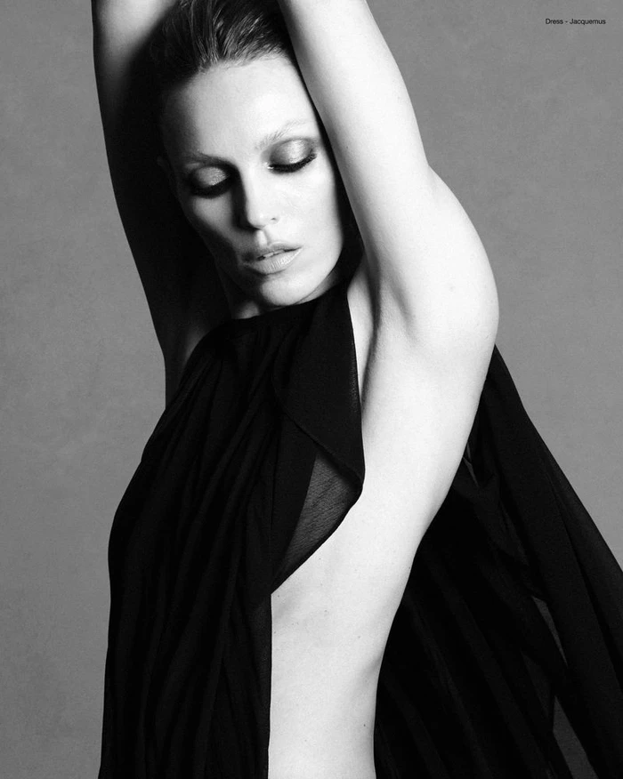 Anja Rubik wearing a black dress with arms raised against a plain neutral background in a black and white portrait.
