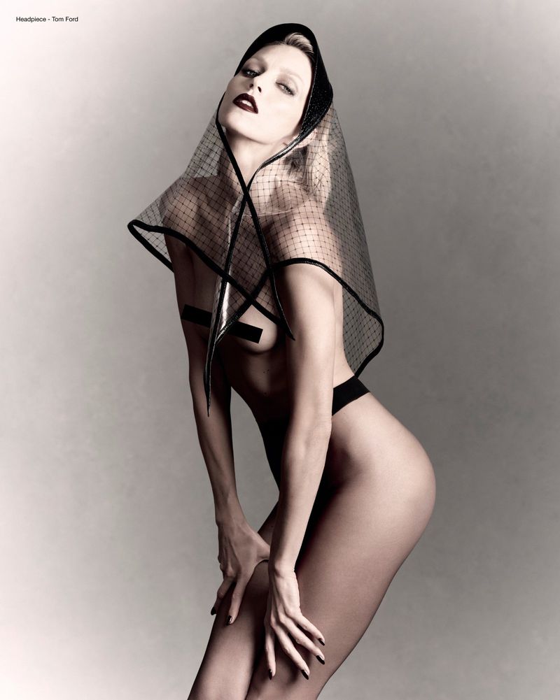 Anja Rubik wearing a mesh headpiece and dark lipstick against a neutral grey studio backdrop.
