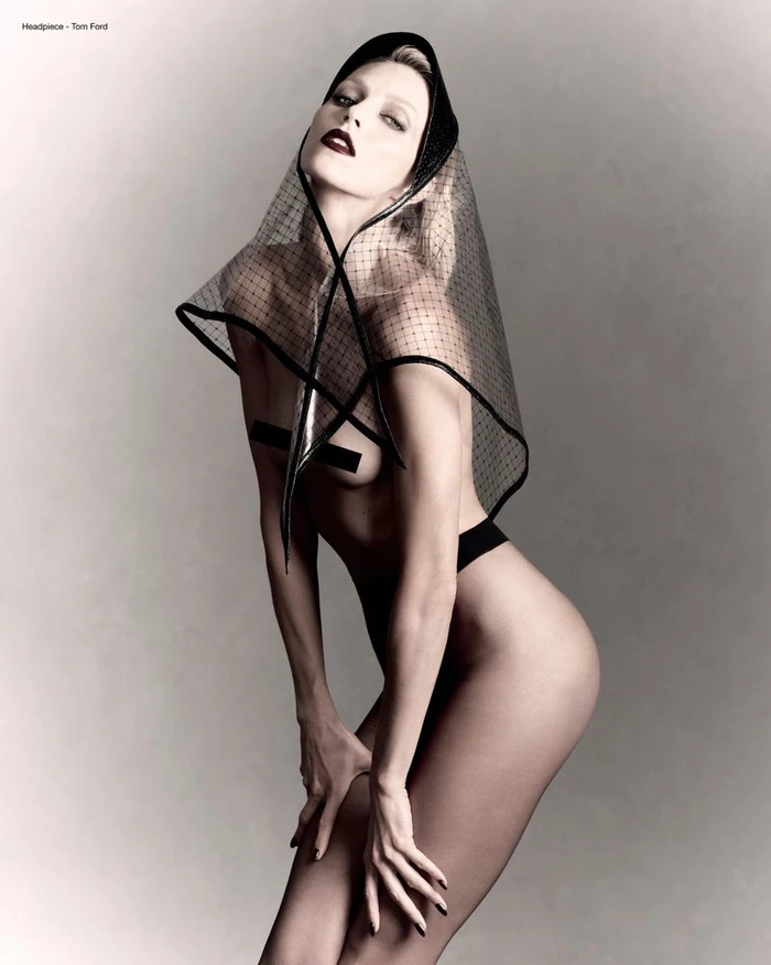 Anja Rubik wearing a mesh headpiece and dark lipstick against a neutral grey studio backdrop.