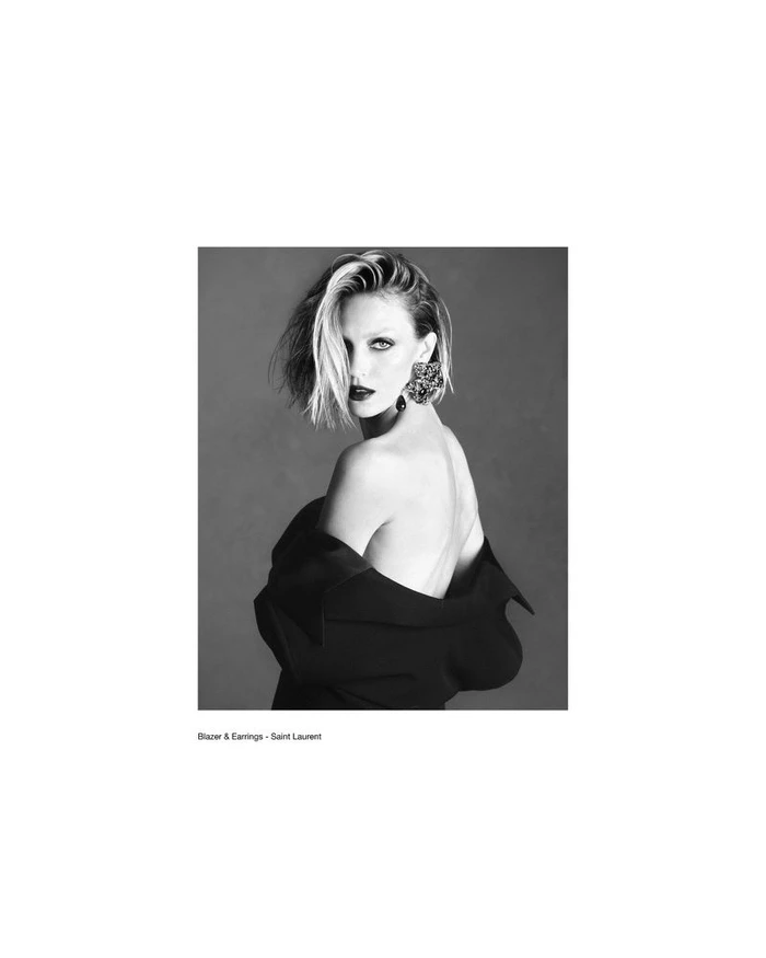 Black and white portrait of Anja Rubik wearing a dark blazer and statement earring, looking back over bare shoulder