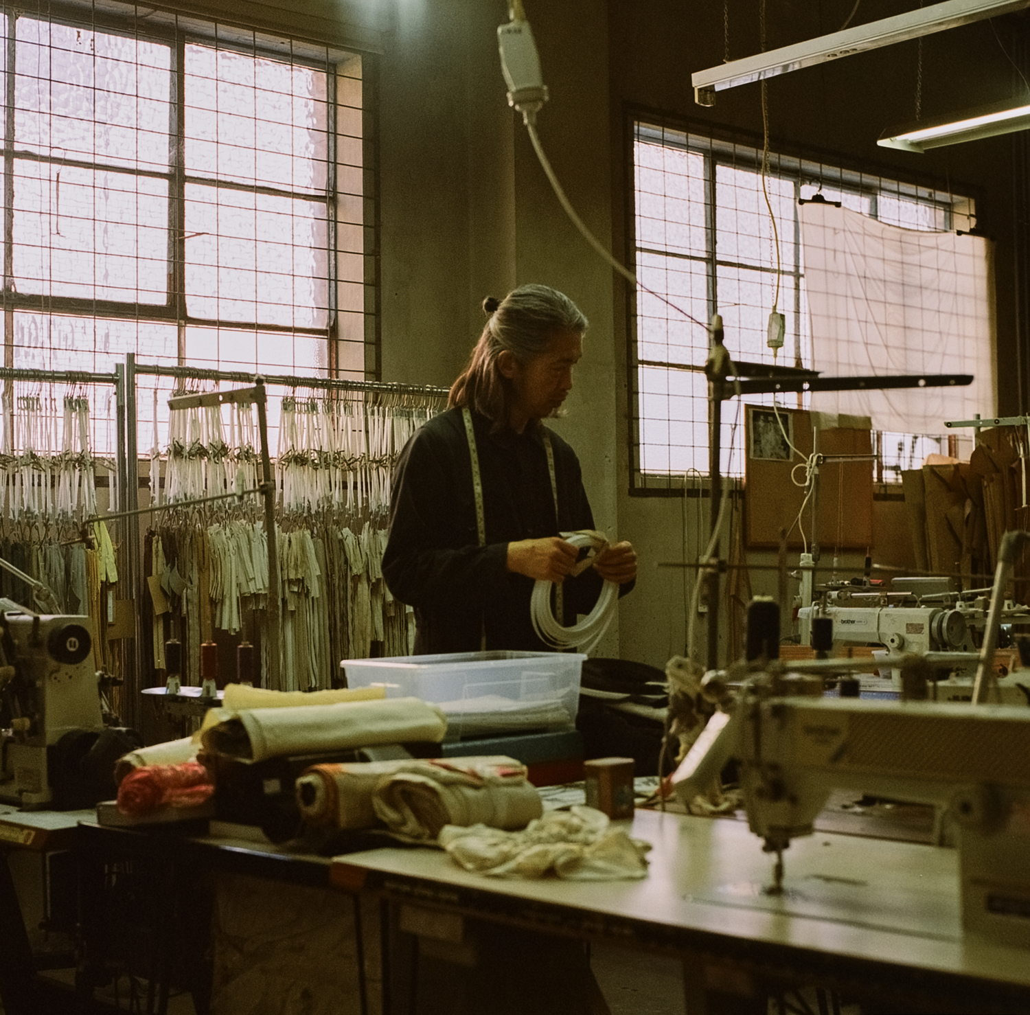 A tailor stands in a bright workshop, holding a measuring tape, with sewing machines, materials, and clothes racks visible in the workspace.