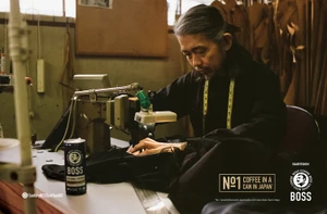 A man with long grey hair seated at a sewing machine working on fabric with a can of Boss coffee on the table.