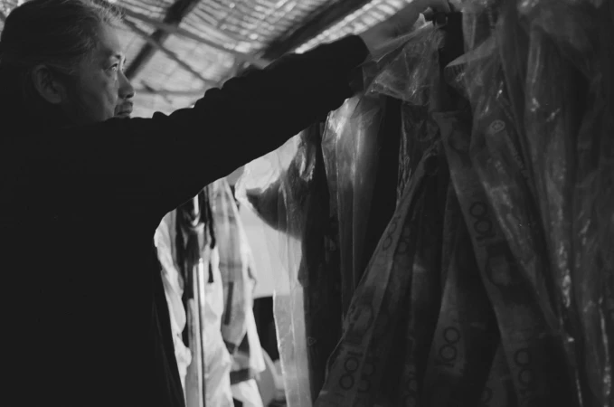 Man reaching for garments wrapped in plastic covers inside a workshop shot in monochrome
