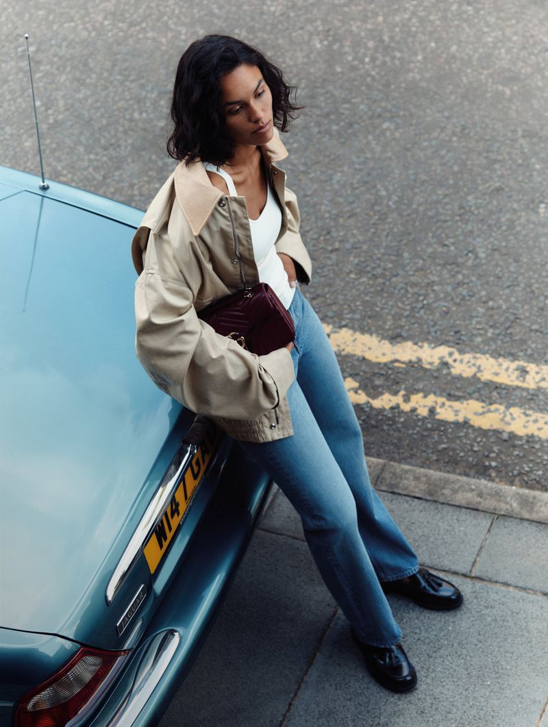 Fashionable adult female leans against a blue car on a city street, wearing a trench coat, jeans, and black shoes, holding a burgundy bag.