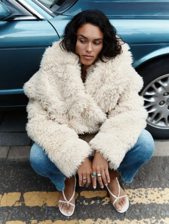 A woman wearing a cream-colored jacket and jeans is crouched on a street with a car in the background, showcasing fashion and urban style.