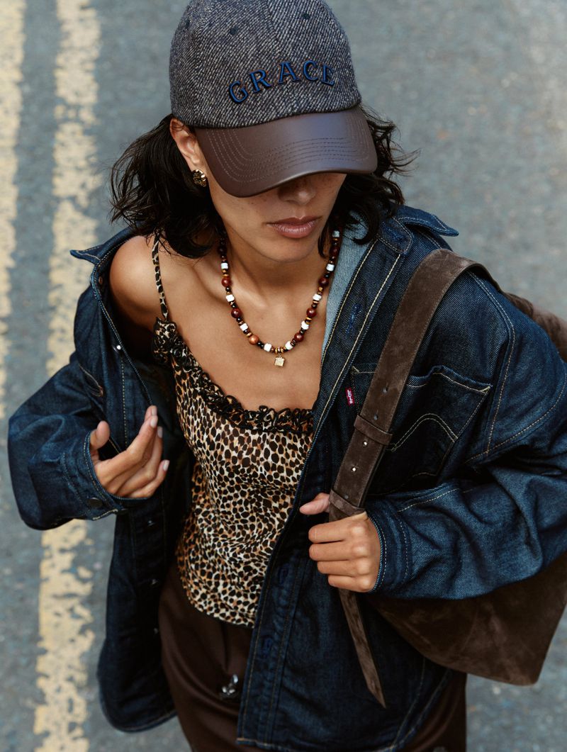 Woman in leopard-print top, denim jacket, and baseball cap with 'Grace' walks on the street holding a brown suede shoulder bag.