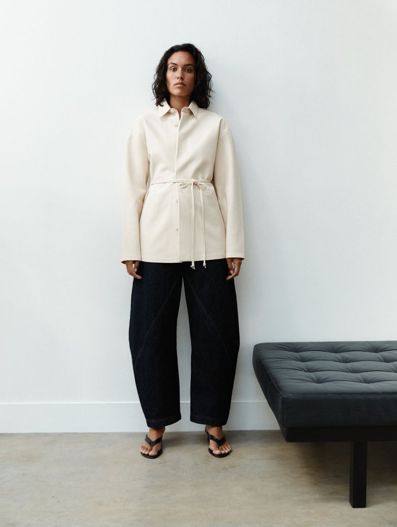 Woman in a beige shirt-jacket with a tie waist and dark blue pants standing indoors against a white wall next to a grey ottoman.