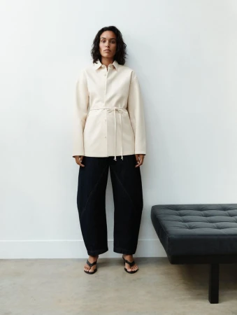 Woman in a beige shirt-jacket with a tie waist and dark blue pants standing indoors against a white wall next to a grey ottoman.