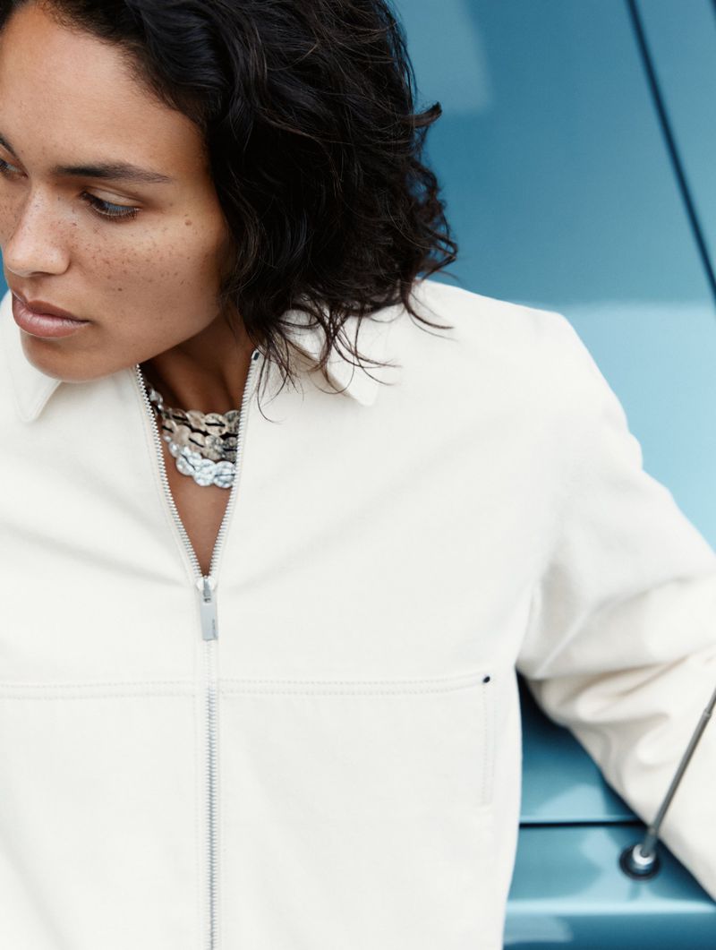 Attractive woman in casual white jacket and jewelry posing near the hood of a classic blue car in a close-up fashion shot.