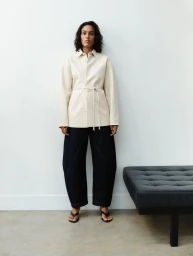 Model wearing a cream button-down shirt and dark denim trousers standing against a plain wall beside a black leather bench.