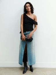 Model standing against a white wall wearing a black one-shoulder top, denim midi skirt, and black boots, holding a clutch.