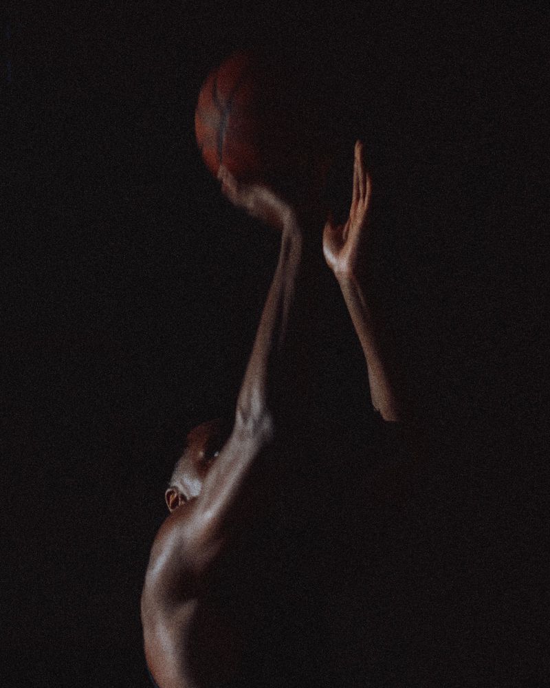 Close-up of a basketball player holding a basketball aloft against a dark background, photographed for V Man.