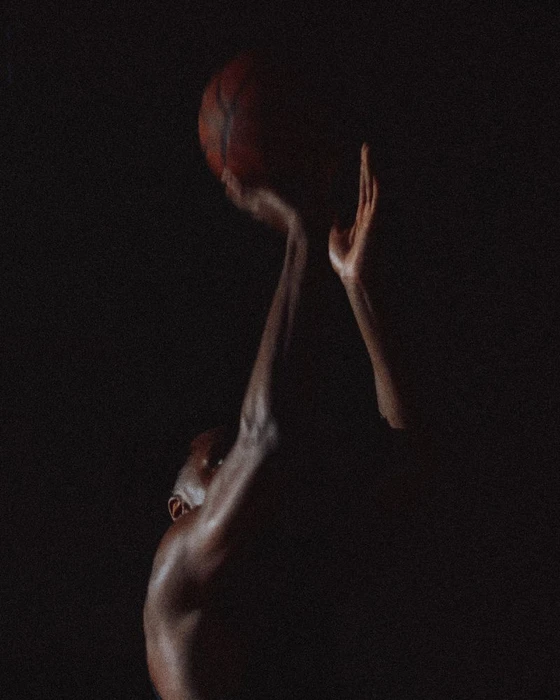 Close-up of a basketball player holding a basketball aloft against a dark background, photographed for V Man.