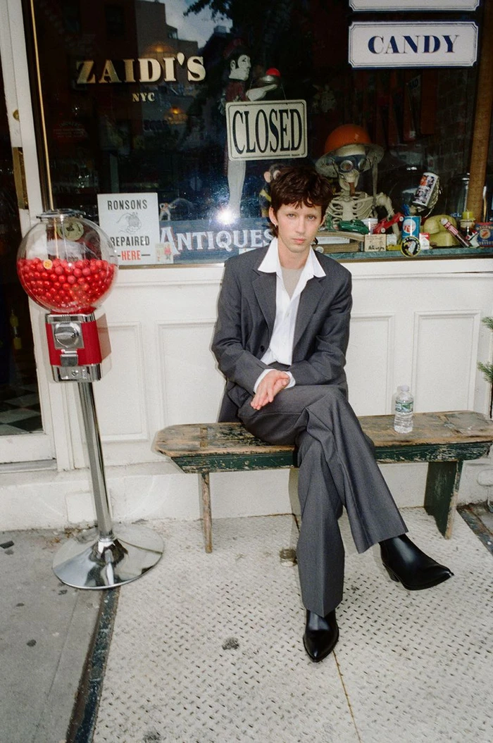 Troye Sivan seated on a wooden bench in a grey suit outside a storefront with a red gumball machine for W Magazine.