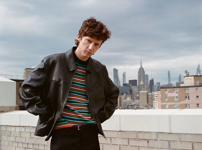 Troye Sivan wearing a black leather jacket and striped sweater on a city rooftop with a skyline background