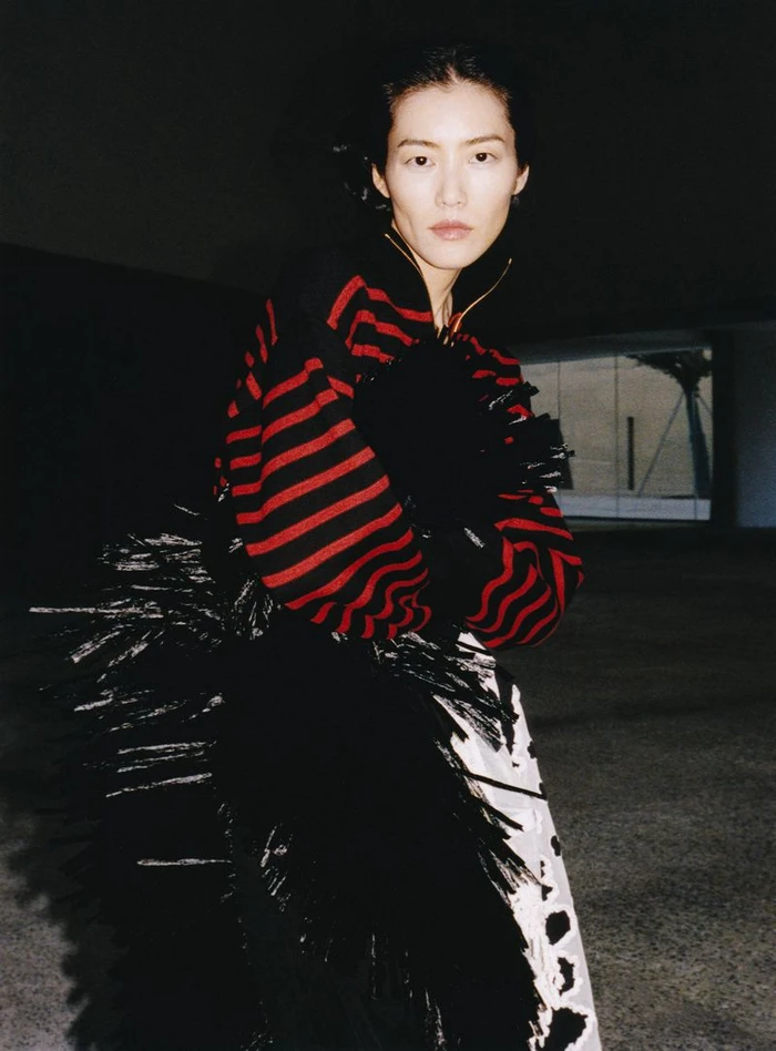 Model Liu Wen in a red and black striped sweater and feathered skirt against a dark architectural background.