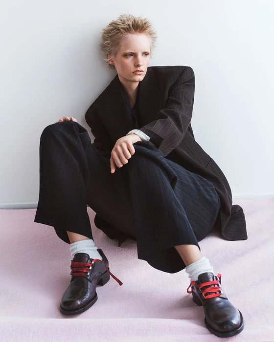 Model with short spiky hair wearing dark pinstripe blazer, trousers, white socks, and leather shoes with red laces