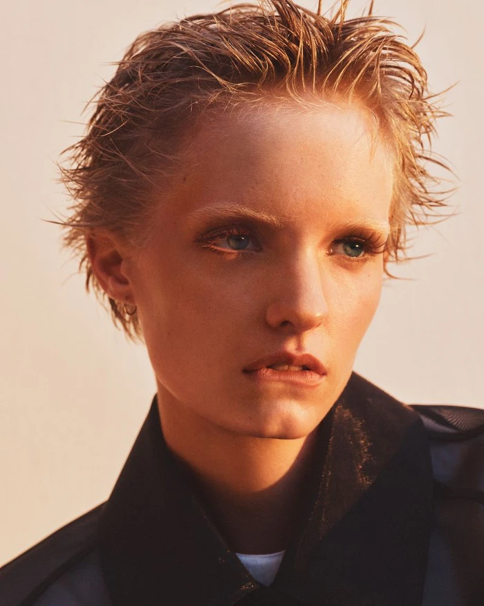Close-up portrait of a model with short wet-look blonde hair wearing a dark collar against a warm plain background.