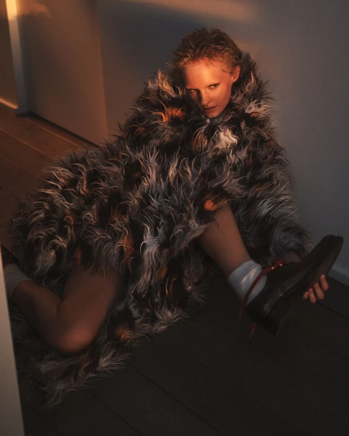 Model Gretchen Easterwood wearing a textured fluffy coat and holding a shoe while seated on a dark wooden floor.