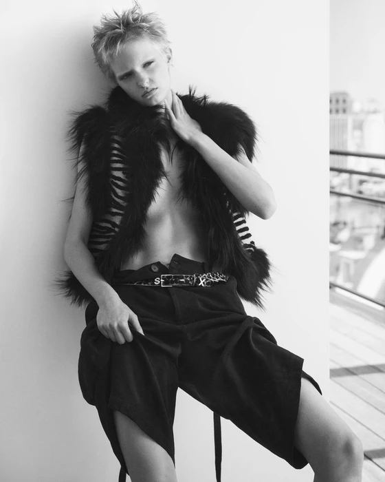 Model Gretchen Easterwood in dark trousers and furry vest leaning against a wall in a black and white editorial by Ilya Lipkin