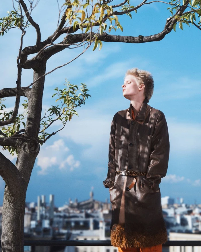 Model with short blond hair wearing a brown coat outdoors against a cityscape and bright blue sky