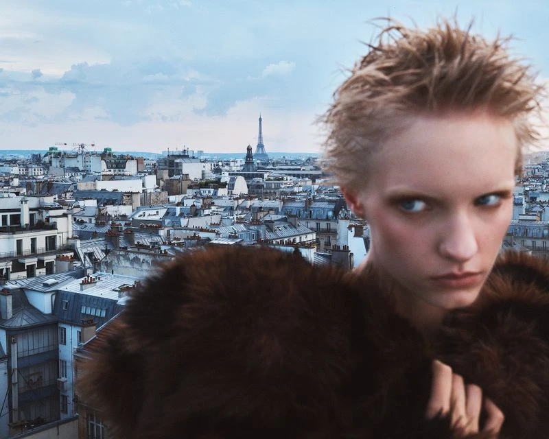 Model with short wet hair and dark faux fur coat standing against a panoramic Parisian rooftop background