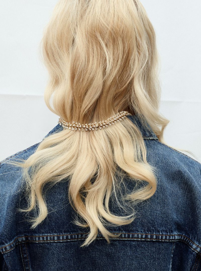 Blonde woman wears a denim jacket with a crystal hair accessory. Hair is shoulder-length. The background is plain white.