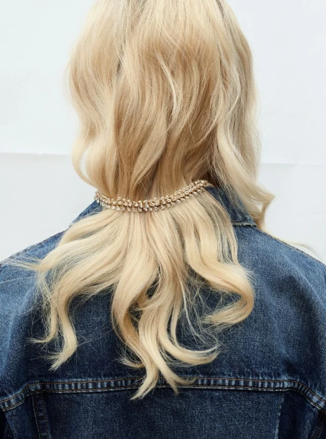 Blonde woman wears a denim jacket with a crystal hair accessory. Hair is shoulder-length. The background is plain white.