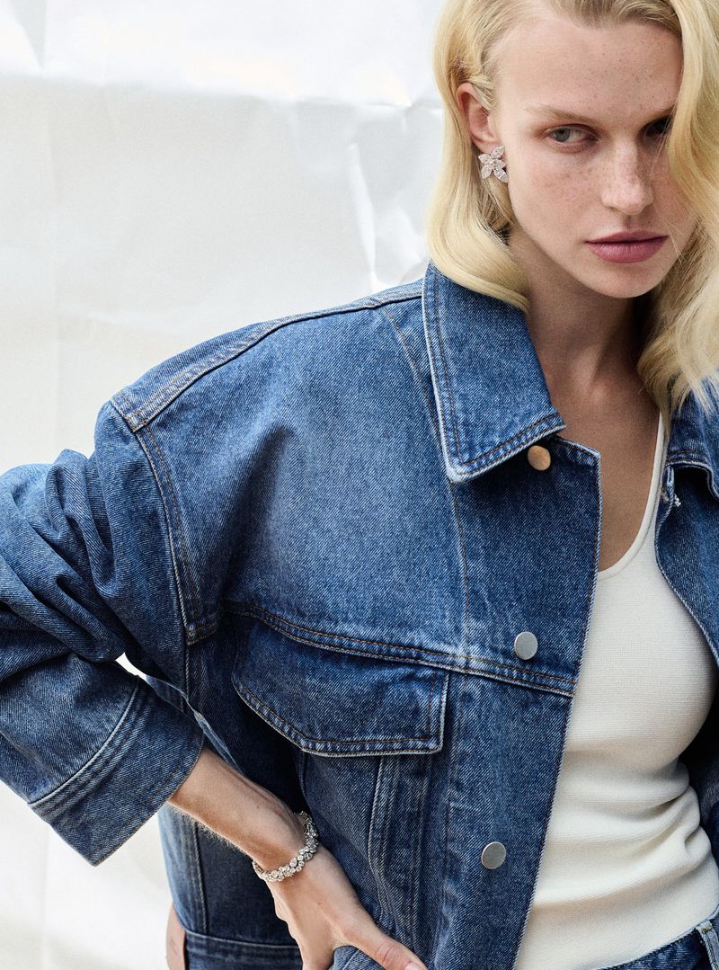 Fashionable woman in denim jacket, jewelry, and light-colored top posing against a bright studio backdrop, showcasing a modern casual style.