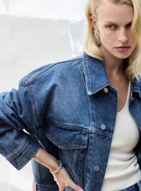 Fashionable woman in denim jacket, jewelry, and light-colored top posing against a bright studio backdrop, showcasing a modern casual style.