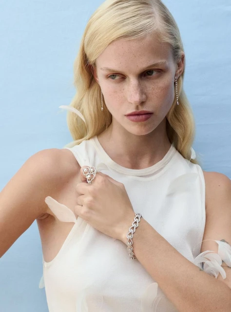 Close-up shot of a fair-skinned woman with blond hair, wearing jewelry and a white dress against a light blue studio backdrop.