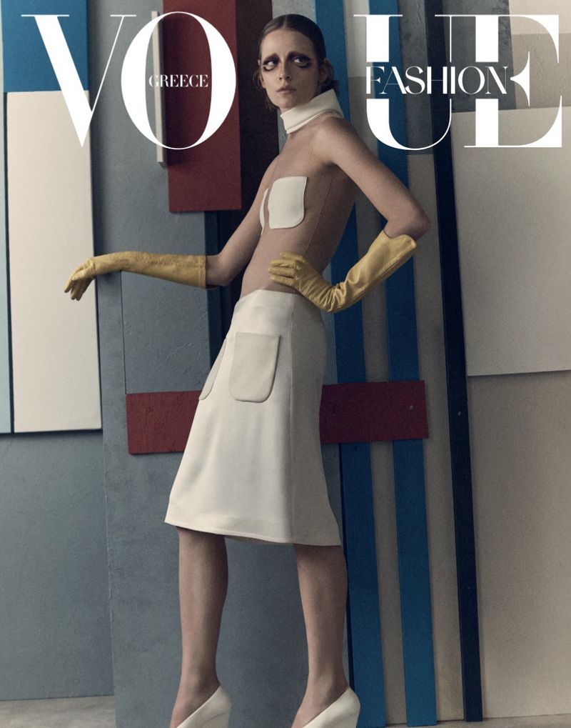 Model wearing a white skirt and yellow gloves posing in front of a geometric panel backdrop for Vogue Greece